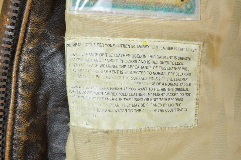 May include: Close-up of a brown leather jacket with a zipper and a tag with care instructions. The tag reads "Authentic Avirex Old Leather Flight Jacket". The leather has a worn appearance.