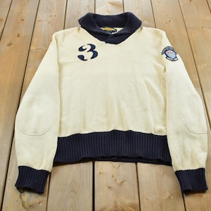 May include: A cream-colored long-sleeve sweater featuring a dark navy blue shawl collar, cuffs, and hem. The front displays a large navy blue number 3 on the right chest and a circular embroidered patch on the left shoulder with elbow patches.
