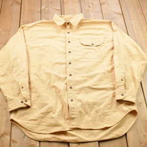 May include: A long-sleeved, button-down shirt in a light yellow colour. The shirt has a single chest pocket and a curved hem.