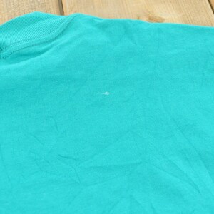 May include: A solid teal t-shirt featuring a ribbed crew neckline and short sleeves. The fabric has a visible texture with a small, faint white mark located near the center of the chest area, showing the plain construction of the garment.