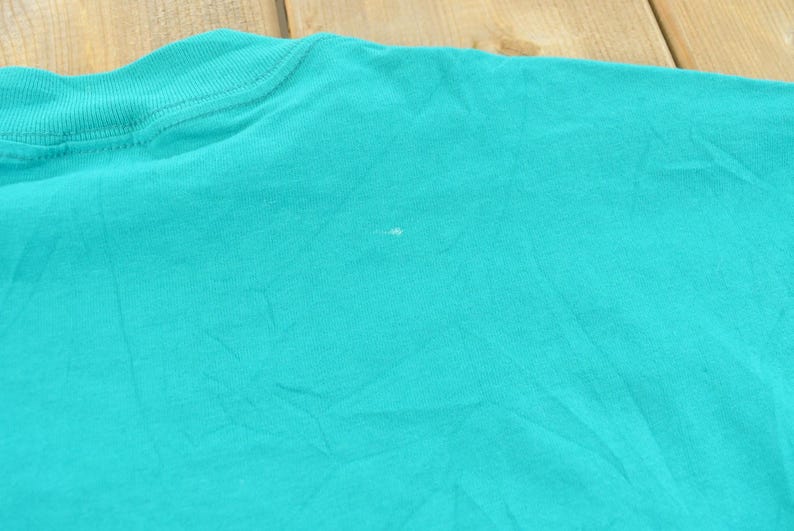 May include: A solid teal t-shirt featuring a ribbed crew neckline and short sleeves. The fabric has a visible texture with a small, faint white mark located near the center of the chest area, showing the plain construction of the garment.