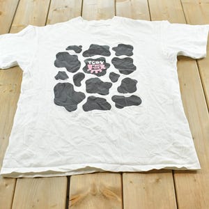 以下が含まれることがあります： 胸元に黒い牛柄の模様が入った白い半袖Tシャツ。プリントの中央には、黒い文字で「TCBY MOO MALT」と書かれた小さなピンク色のグラフィックがあります。このシャツはクルーネックで、裾はストレートなデザインです。