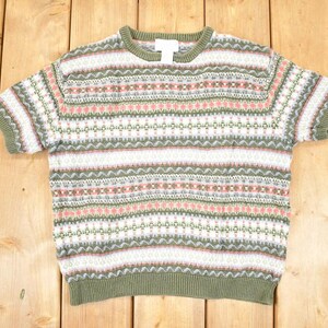 Könnte beinhalten: Kurzärmliger Pullover mit einem gemusterten Design in Grün-, Weiß- und Rosatönen. Der Pullover hat einen Rundhalsausschnitt und gerippte Bündchen und Saum. Das Kleidungsstück ist aus Strickstoff gefertigt und wird auf einer Holzoberfläche präsentiert.