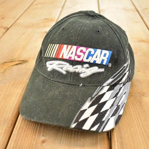 Può includere: Un cappellino da baseball nero NASCAR Racing. Il cappellino presenta il logo NASCAR in bianco, rosso, blu e giallo. Il lato del cappellino ha un motivo a bandiera a scacchi in bianco e nero.
