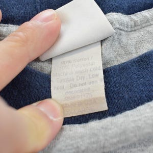 Puede incluir: Primer plano de una camiseta gris y azul marino con una etiqueta blanca. La etiqueta tiene el texto "90% Algod&oacute;n / 10% Poli&eacute;ster. Lavar a m&aacute;quina en fr&iacute;o. Secar en secadora, a baja temperatura. No planchar. decorado. RN 91173."