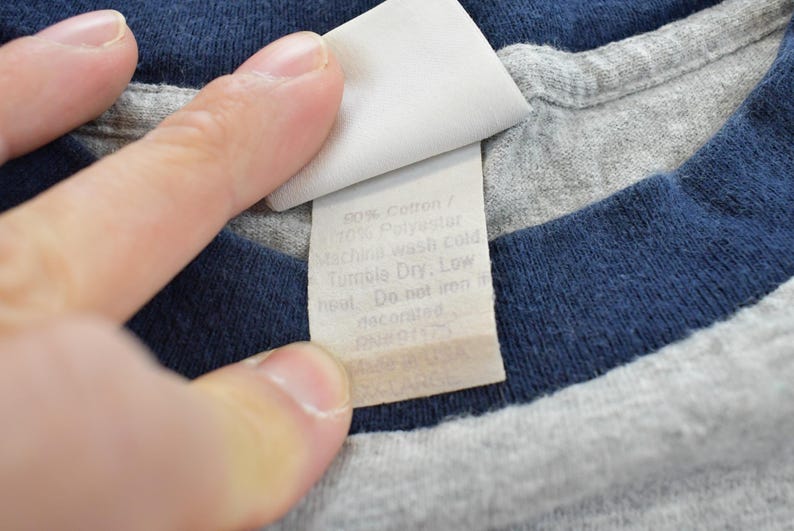 Puede incluir: Primer plano de una camiseta gris y azul marino con una etiqueta blanca. La etiqueta tiene el texto "90% Algod&oacute;n / 10% Poli&eacute;ster. Lavar a m&aacute;quina en fr&iacute;o. Secar en secadora, a baja temperatura. No planchar. decorado. RN 91173."