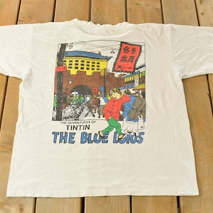 May include: White t-shirt featuring a colorful graphic from "The Adventures of Tintin: The Blue Lotus." The design depicts a street scene with a figure in a red jacket and green pants. The text "THE BLUE LOTUS" is printed below the image.