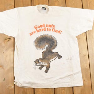May include: White t-shirt featuring a cartoon squirrel and the orange text, "Good nuts are hard to find!" The squirrel is brown and grey with a large, fluffy tail. The shirt is displayed on a wooden surface.
