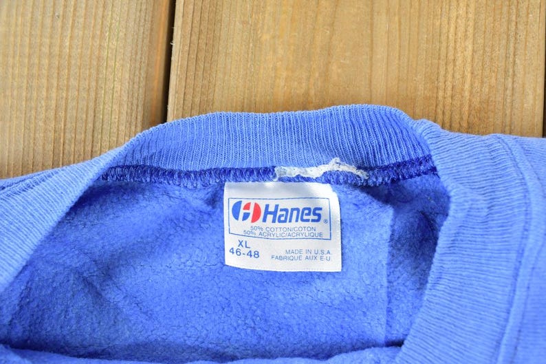 Puede incluir: Una sudadera azul Hanes con una etiqueta que dice "30% Algod&oacute;n/Coton 50% Acr&iacute;lico/Acrylique" y "XL 46-48" y "Hecho en EE. UU. Fabrique aux E.U."