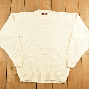 May include: A cream-colored knitted sweater with a cable knit pattern. The sweater has a crew neck and long sleeves.