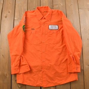 Puede incluir: Camisa de trabajo naranja de manga larga con un parche blanco que dice "Waukesha Gun Club Hunter Safety" y un parche verde en la manga izquierda. La camisa tiene un nombre "Jake" bordado en la parte delantera.