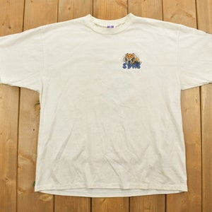 Puede incluir: Camiseta blanca con un gráfico de tigre de dibujos animados y la palabra "SDUHS" impresa en azul.