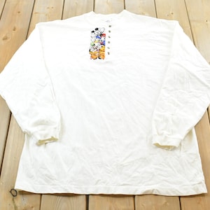 May include: White long-sleeved shirt featuring a graphic of cartoon characters stacked vertically on the upper chest. The shirt has a crew neck and buttons down the front. The sleeves have ribbed cuffs.