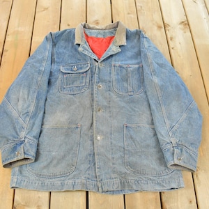 May include: A light blue denim jacket with a corduroy collar and a red interior lining. The jacket has four front pockets and button closures. The jacket is laid flat on a wooden surface.
