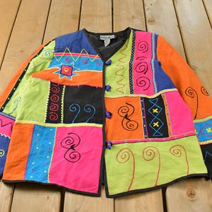May include: A long-sleeved jacket featuring a vibrant patchwork design in bright orange, lime green, hot pink, blue, and black. The garment includes various embroidered patterns such as spirals, zigzags, and geometric shapes, with three toggle closures.