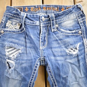 K&ouml;nnte beinhalten: Blaue Jeans aus Denim mit Destroyed-Look, wei&szlig;en N&auml;hten und Verzierungen. Die Jeans haben ein zerrissenes Design mit einem gestreiften Stoff-Patch am linken Bein. Der Bund tr&auml;gt den Schriftzug "Rock Revival" in Orange.