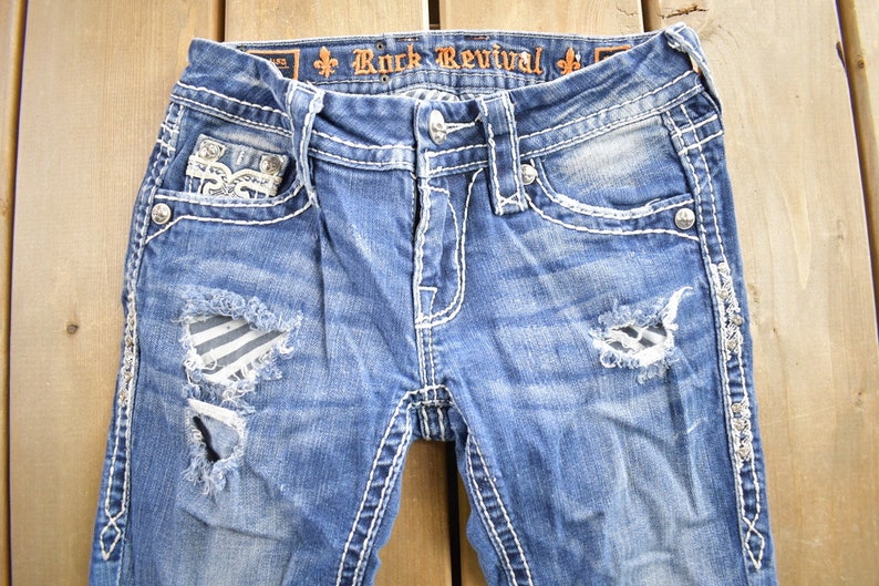 K&ouml;nnte beinhalten: Blaue Jeans aus Denim mit Destroyed-Look, wei&szlig;en N&auml;hten und Verzierungen. Die Jeans haben ein zerrissenes Design mit einem gestreiften Stoff-Patch am linken Bein. Der Bund tr&auml;gt den Schriftzug "Rock Revival" in Orange.