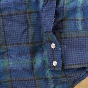 May include: Detail of a blue, green, and black plaid flannel shirt. The cuff is buttoned with two silver buttons. The fabric has a textured look. The shirt's pattern and style suggest a vintage or classic design.