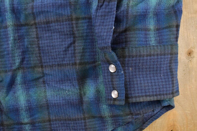 May include: Detail of a blue, green, and black plaid flannel shirt. The cuff is buttoned with two silver buttons. The fabric has a textured look. The shirt's pattern and style suggest a vintage or classic design.