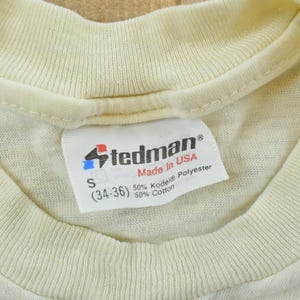 May include: A cream-colored t-shirt featuring a white interior neck label with the Stedman logo, Made in USA text, size S (34-36), and a fabric composition of 50% Kodel polyester and 50% cotton. The garment has a ribbed crew neckline.
