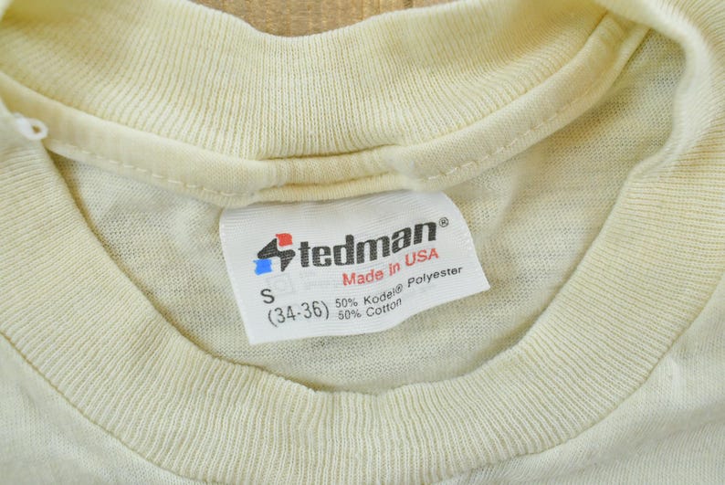 May include: A cream-colored t-shirt featuring a white interior neck label with the Stedman logo, Made in USA text, size S (34-36), and a fabric composition of 50% Kodel polyester and 50% cotton. The garment has a ribbed crew neckline.