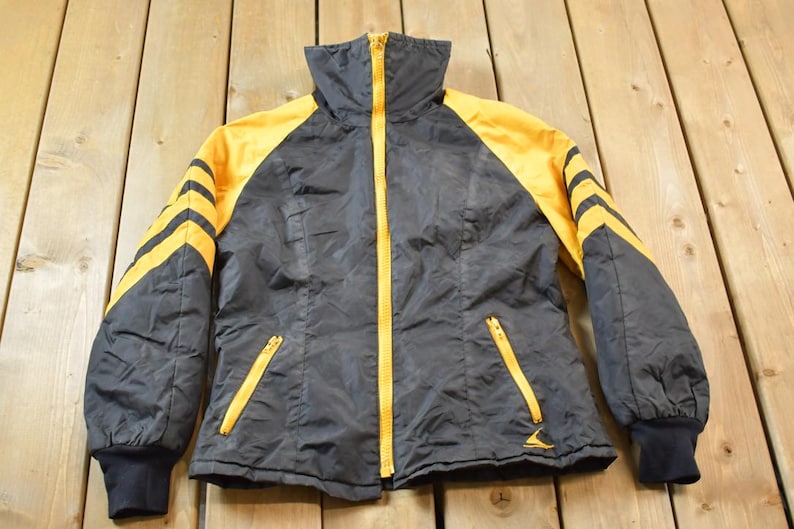 May include: A black and yellow jacket with a zipper closure and a high collar. The jacket has yellow stripes on the sleeves and a yellow zipper.