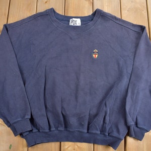 May include: A navy blue sweatshirt with a small embroidered logo on the left chest. The logo features a shield with a crown and the text "PDX" inside.