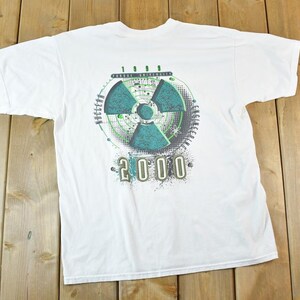May include: White t-shirt with a large graphic on the front. The graphic features a teal and green radiation symbol inside a circle with the text "1999" above and "2000" below. The shirt is laid flat on a wooden surface.