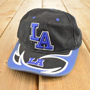 Op de afbeelding: Zwarte baseballcap met een blauwe klep en de letters "LA" in blauw en wit. De cap heeft een gebogen klep met een gestileerd ontwerp en de letters "LA" in blauw.