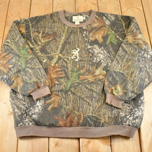 May include: A camouflage-patterned sweatshirt with a crew neck and long sleeves. The design features brown, green, and grey tones with leaf and branch details. A white Browning logo is centered on the chest. The cuffs and hem are a solid brown.