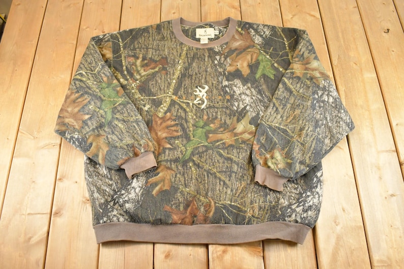 May include: A camouflage-patterned sweatshirt with a crew neck and long sleeves. The design features brown, green, and grey tones with leaf and branch details. A white Browning logo is centered on the chest. The cuffs and hem are a solid brown.