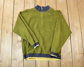Könnte beinhalten: Ein grüner Fleece-Pullover mit einem halben Reißverschluss und einem dunkelblauen und gelben Saum an Hals und Bund.