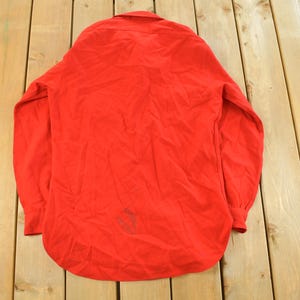 May include: A bright red long-sleeve shirt with a button-down collar. The shirt is made of a soft material and has a curved hem. The sleeves have buttoned cuffs. The shirt is laid out on a wooden surface.