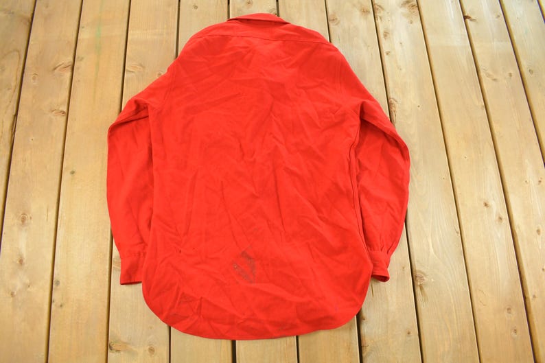 May include: A bright red long-sleeve shirt with a button-down collar. The shirt is made of a soft material and has a curved hem. The sleeves have buttoned cuffs. The shirt is laid out on a wooden surface.