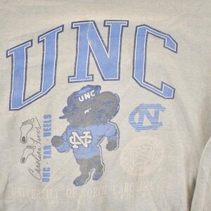 K&ouml;nnte beinhalten: Hellgraues Sweatshirt mit den Buchstaben "UNC" in Hellblau und einer Cartoon-Tar Heel-Maskottchen-Grafik. Zus&auml;tzlicher Text enth&auml;lt "Carolina Tar Heels" und ein Universit&auml;tslogo. Ein Vintage-College-Kleidungsst&uuml;ck.