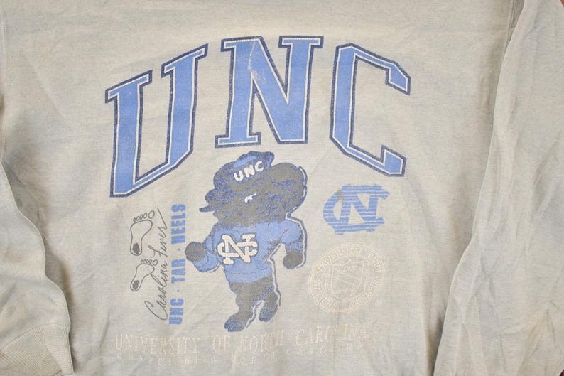 K&ouml;nnte beinhalten: Hellgraues Sweatshirt mit den Buchstaben "UNC" in Hellblau und einer Cartoon-Tar Heel-Maskottchen-Grafik. Zus&auml;tzlicher Text enth&auml;lt "Carolina Tar Heels" und ein Universit&auml;tslogo. Ein Vintage-College-Kleidungsst&uuml;ck.