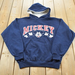 K&ouml;nnte beinhalten: Marineblauer Kapuzenpullover mit wei&szlig;em Innenfutter. Auf der Vorderseite steht das Wort "MICKEY" in Rot und Wei&szlig;, mit Mickey Mouses Gesicht und Silhouette. Ein klassischer, bequemer Hoodie.