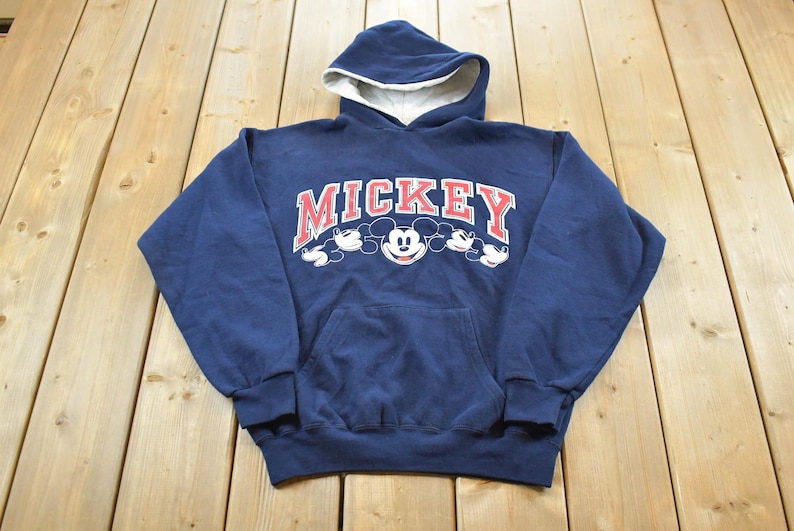 K&ouml;nnte beinhalten: Marineblauer Kapuzenpullover mit wei&szlig;em Innenfutter. Auf der Vorderseite steht das Wort "MICKEY" in Rot und Wei&szlig;, mit Mickey Mouses Gesicht und Silhouette. Ein klassischer, bequemer Hoodie.