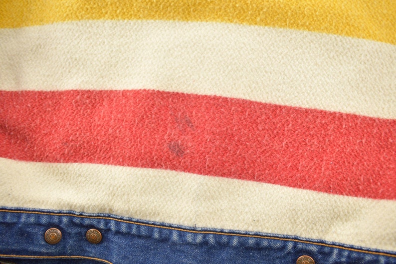 May include: Close-up of a blue denim jacket with a red, white, and yellow striped flannel lining. The jacket has two metal buttons.