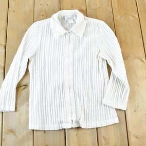 May include: White, long-sleeved shirt with a ribbed texture and a collared neckline. The shirt has a full-length zipper and is made of a lightweight fabric. The garment is displayed on a wooden surface.