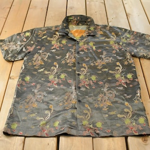 May include: A short-sleeved button-down shirt in a dark charcoal gray fabric featuring an intricate woven pattern of golden and red dragons, clouds, and floral motifs. The shirt has a classic collar, a front button closure, and a chest pocket.