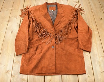 May include: A brown suede jacket with fringe detailing around the collar and front. The jacket has a button closure and two pockets.