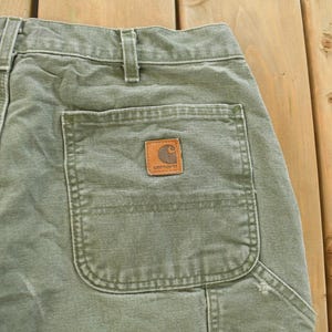 Puede incluir: Primer plano de la parte trasera de unos pantalones de trabajo verde oliva. Los pantalones tienen un bolsillo trasero grande con un parche marr&oacute;n de la marca Carhartt. Los pantalones est&aacute;n hechos de una tela duradera y tienen costuras visibles.