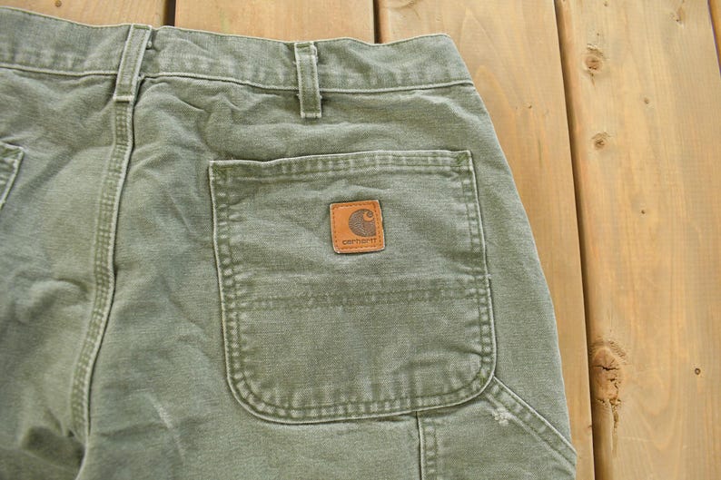 Puede incluir: Primer plano de la parte trasera de unos pantalones de trabajo verde oliva. Los pantalones tienen un bolsillo trasero grande con un parche marr&oacute;n de la marca Carhartt. Los pantalones est&aacute;n hechos de una tela duradera y tienen costuras visibles.