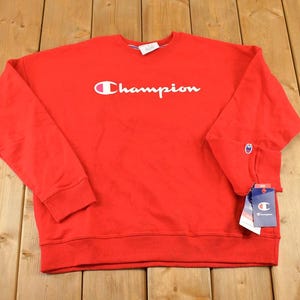 Könnte beinhalten: Ein leuchtend rotes Langarm-Sweatshirt mit dem weißen Champion-Markenlogo auf der Brust. Das Kleidungsstück verfügt über einen gerippten Rundhalsausschnitt, gerippte Bündchen und einen gerippten Saum. Ein kleines Champion-Logo-Patch ist am linken Ärmel sichtbar.