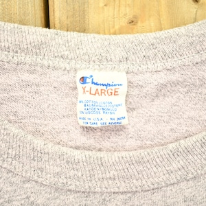 May include: A gray Champion brand t-shirt tag with the text "X-Large" and the fabric content: "91% Cotton/Coton, Baumwoll/Cotone, Katoen/Bomuld, 9% Viscose Rayon". The tag also says "Made in U.S.A. RN 26094" and "For Care See Reverse".