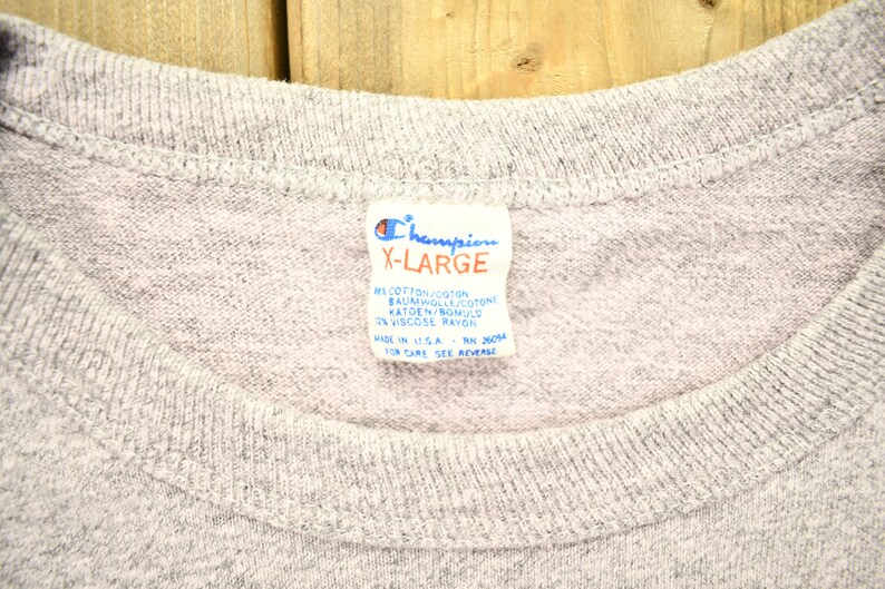 May include: A gray Champion brand t-shirt tag with the text "X-Large" and the fabric content: "91% Cotton/Coton, Baumwoll/Cotone, Katoen/Bomuld, 9% Viscose Rayon". The tag also says "Made in U.S.A. RN 26094" and "For Care See Reverse".