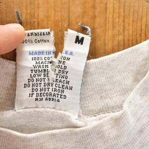 以下が含まれることがあります： 薄いグレーのTシャツのクローズアップ。タグには「OVERSIZED 100% Cotton MADE IN U.S.A.」と洗濯表示が記載されています。シャツにはMサイズのラベルが付いています。