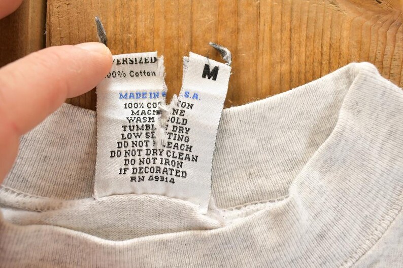 以下が含まれることがあります： 薄いグレーのTシャツのクローズアップ。タグには「OVERSIZED 100% Cotton MADE IN U.S.A.」と洗濯表示が記載されています。シャツにはMサイズのラベルが付いています。