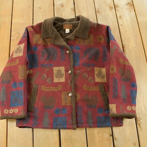 Peut inclure: Veste rouge avec col et bordures marron, ornée d'un motif d'arbres, d'ours et de canoës. La veste a deux poches et est boutonnée. La veste est faite d'une matière douce.